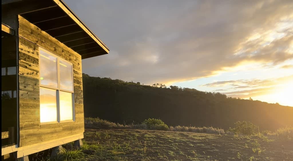 Golden-hour light over Kuahiwi Farms in East Moloka'i: a small wooden shelter on the left and rolling hills under a warm, cloudy sky.